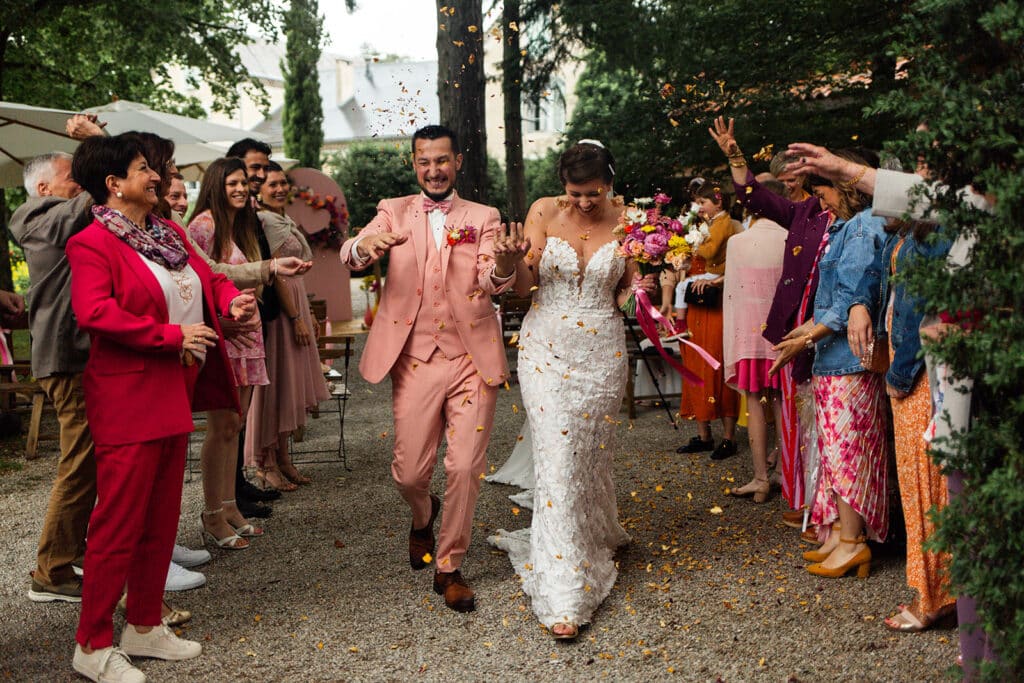 Sortie de cérémonie de mariage d’un couple souriant sous les applaudissements et les confettis des invités en extérieur.