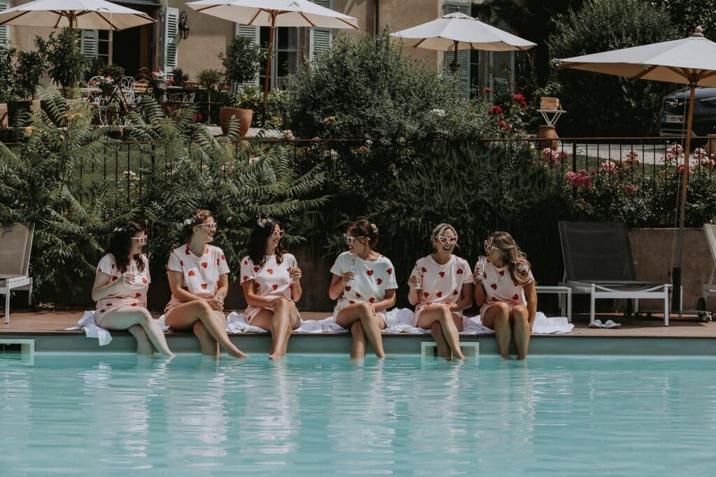 Six femmes en pyjamas roses à cœurs et lunettes fantaisie rient au bord d'une piscine avec du champagne, en été.
