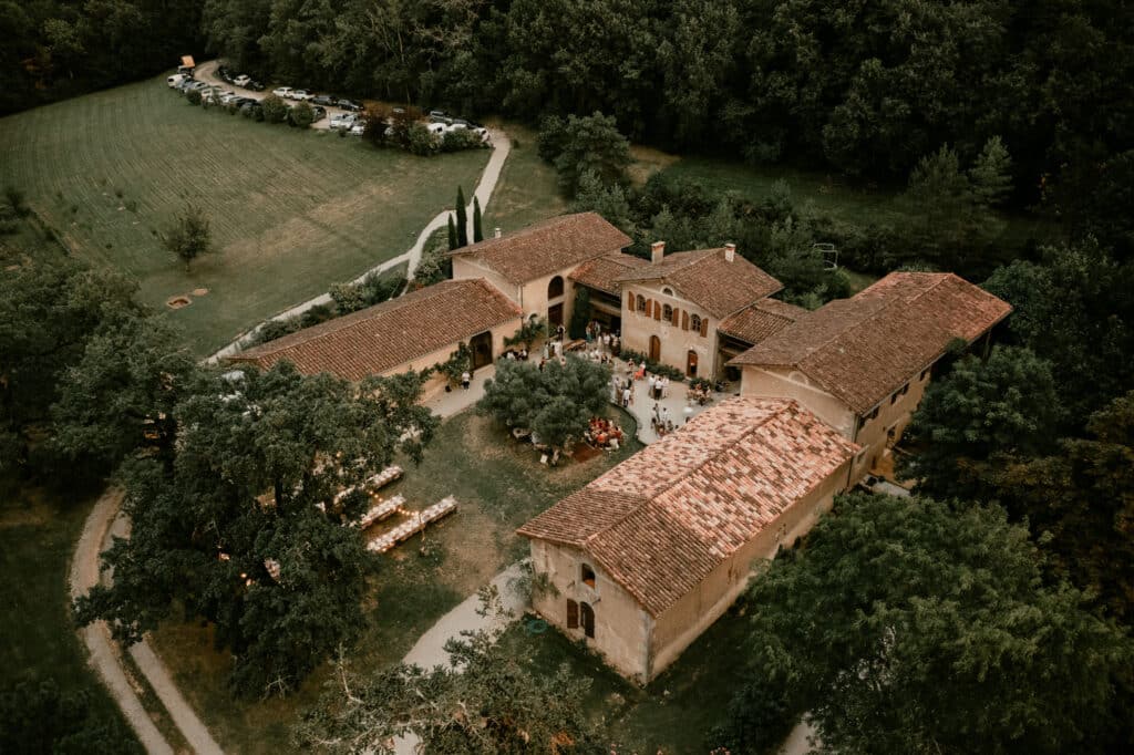 Vue aérienne d'un domaine en pierre avec toits en tuiles, où se déroule une célébration extérieure. Invités et tables illuminées.