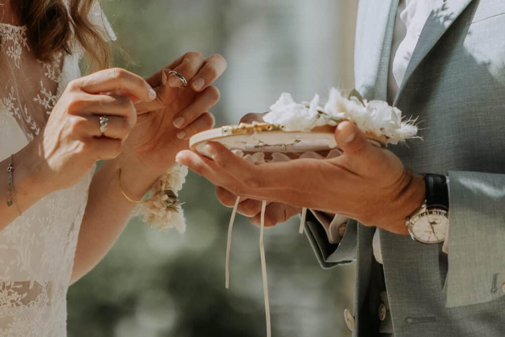 Un couple échange des anneaux de mariage lors d'une cérémonie intime. La femme tient une bague, l'homme présente le porte-alliances fleuri.