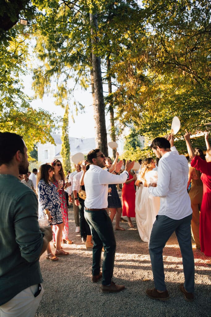 Foule joyeuse dansant en plein air. Deux hommes rient, d'autres tiennent des éventails sous des arbres ensoleillés, près d'un château.