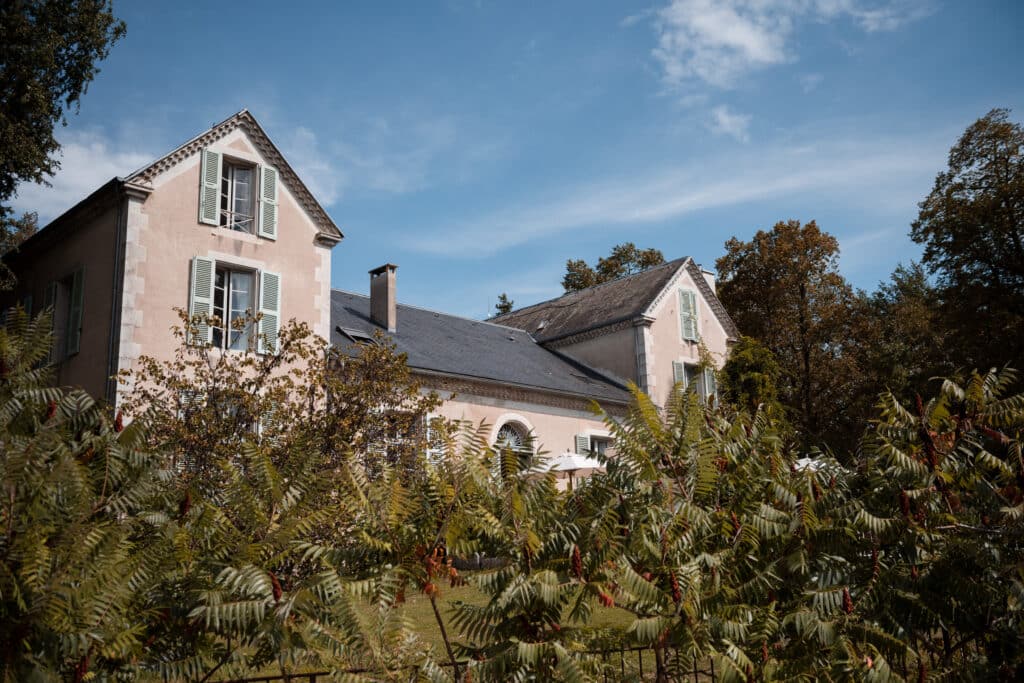 Maison de campagne aux murs clairs et volets verts, entourée d'un jardin luxuriant sous ciel bleu. Bâtiment historique paisible.