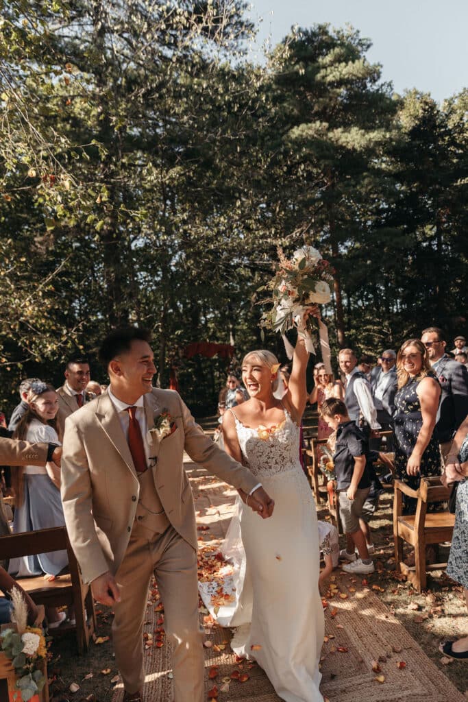 Mariés joyeux descendent l'allée en plein air. La mariée lève son bouquet, des pétales de roses jonchent le sol.