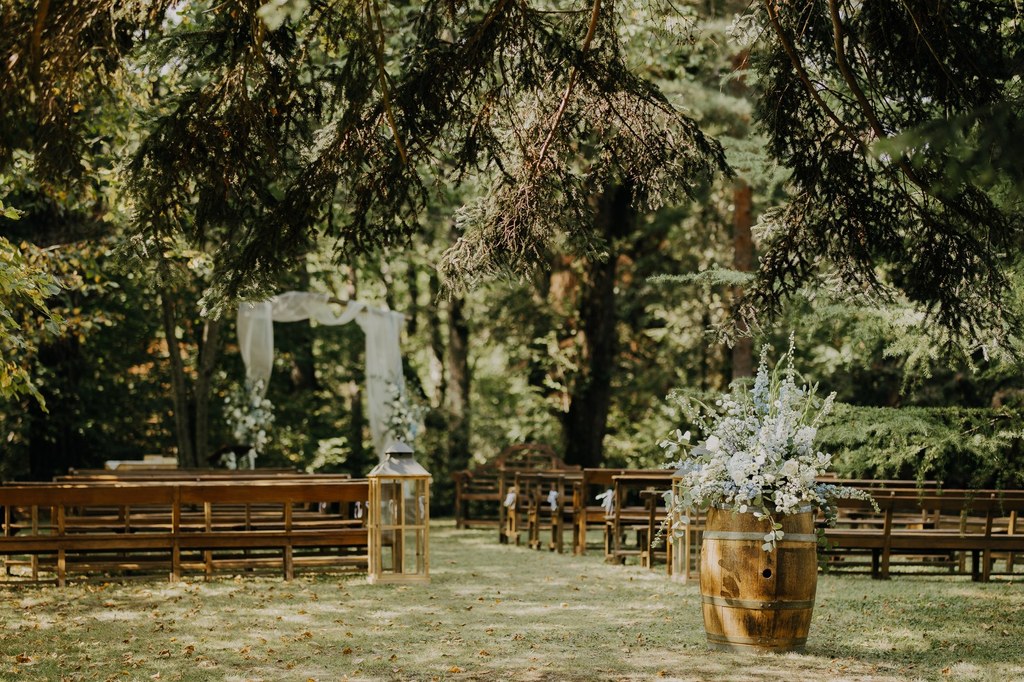 Cérémonie de mariage en plein air dans un cadre boisé. Allée avec arche fleurie blanche, bancs en bois, lanterne et tonneau floral.