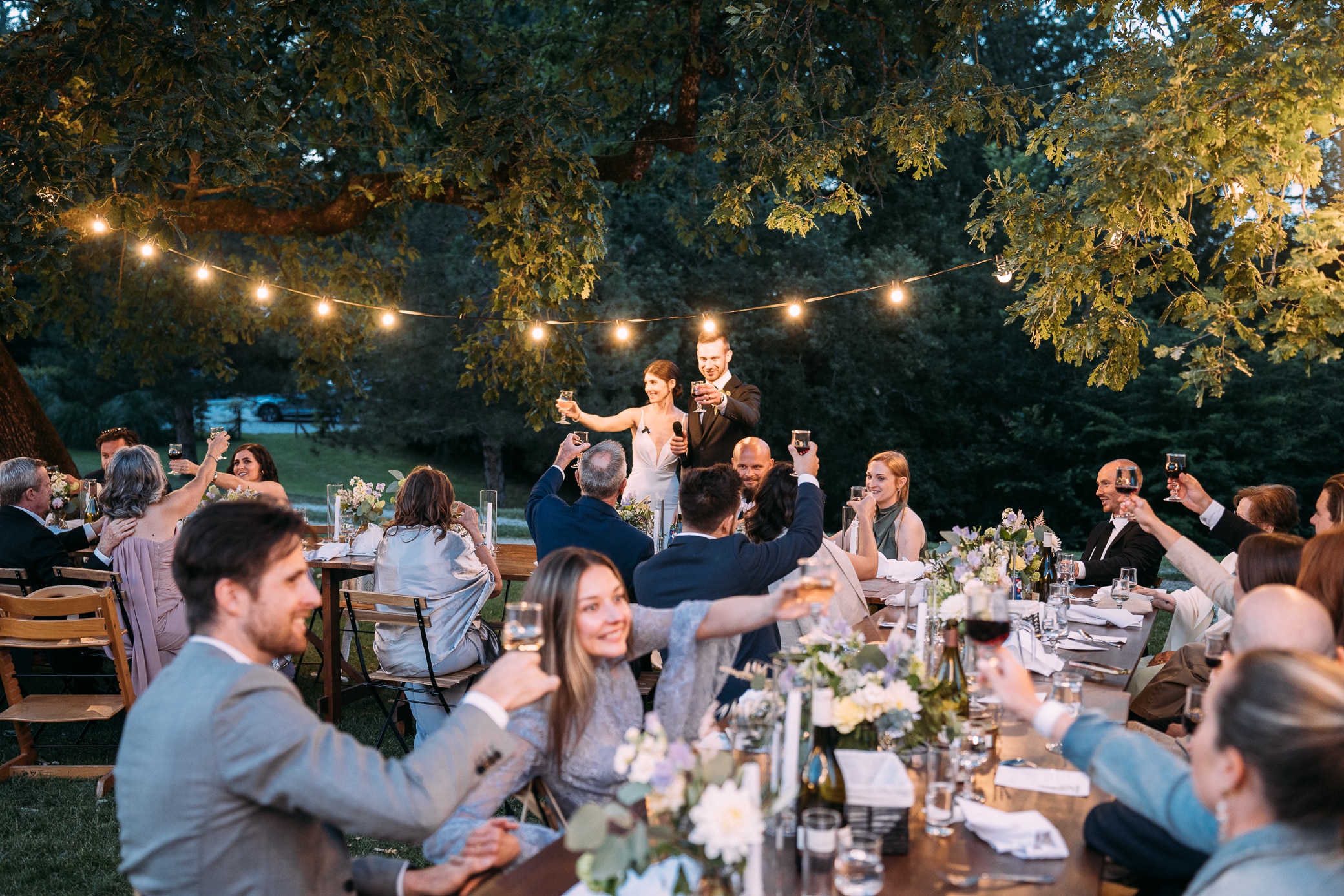 Couple et invités souriants toastant à une réception de mariage en plein air, éclairée par des guirlandes lumineuses.