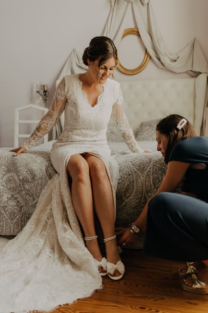 Mariée souriante en robe de dentelle blanche, assise sur un lit. Une femme l'aide à enfiler ses chaussures élégantes à nœuds et perles.
