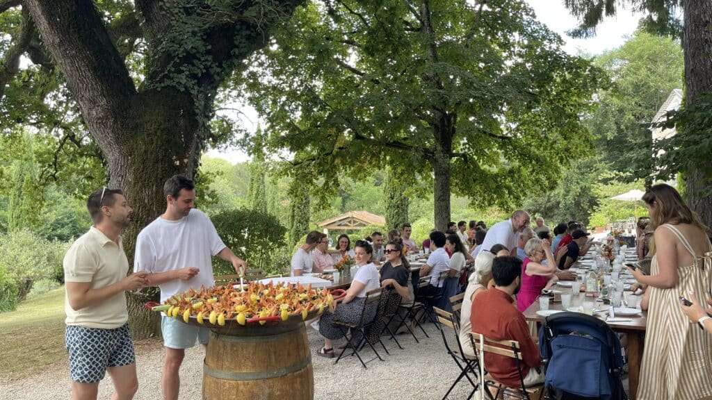 Repas de groupe en extérieur. Les mariés servent une paella géante garnie de crevettes et citron à leurs convives attablés sous les arbres.