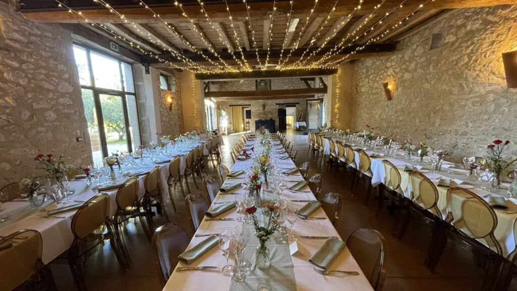 Grande salle de réception rustique avec murs en pierre et plafond boisé orné de guirlandes lumineuses. Longues tables dressées pour un événement avec vaisselle et bouquets de fleurs. Vue sur le jardin par de grandes fenêtres, signe EXIT visible.