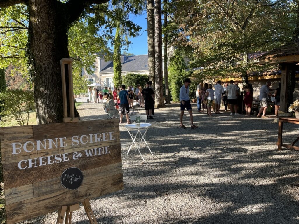 Panneau "BONNE SOIREE CHEESE & WINE" et "Olivia + Robert". Invités dégustant vin et fromage dans les jardins d'un château ensoleillé.