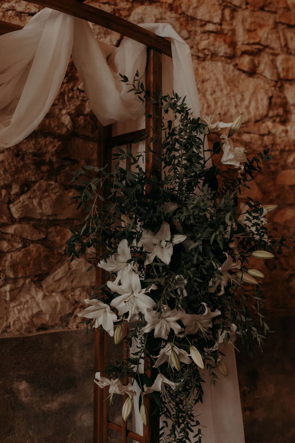 Décoration florale riche en lys blancs et feuillage vert foncé sur une arche en bois rustique devant un mur de pierres ocres.