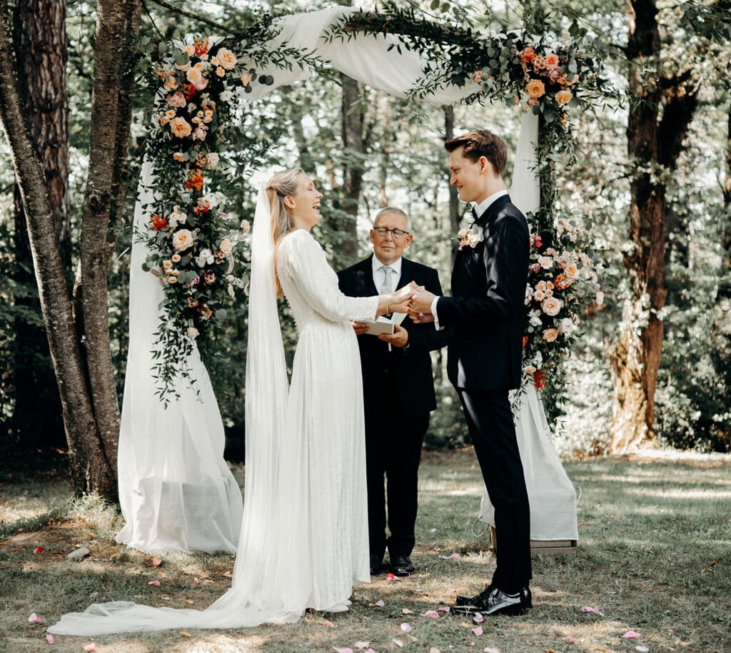 Mariage en plein air sous une arche fleurie : la mariée rit aux éclats en tenant les mains de son marié devant l'officiant.