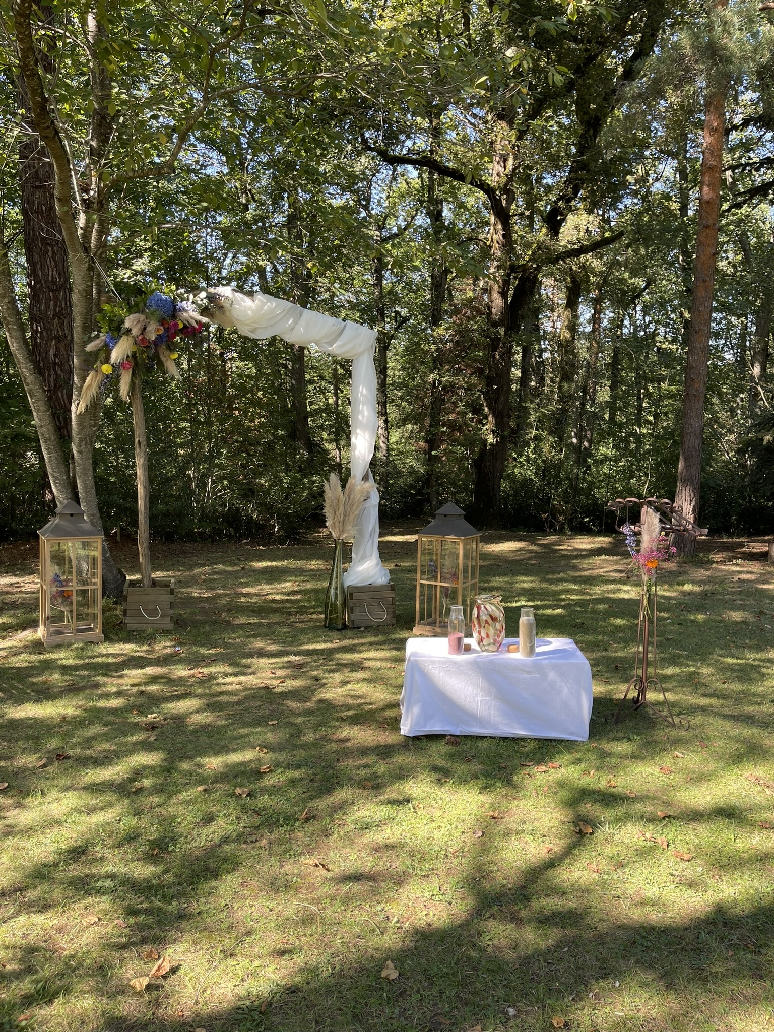 Cérémonie de mariage en plein air sous une arche fleurie, avec table blanche et lanternes décoratives dans un cadre boisé.
