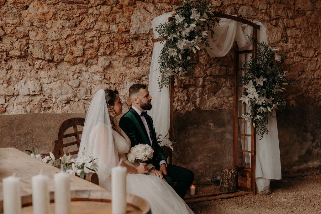 Mariés assis devant une arche décorée de lys blancs et de verdure, sur fond de mur en pierre rustique.