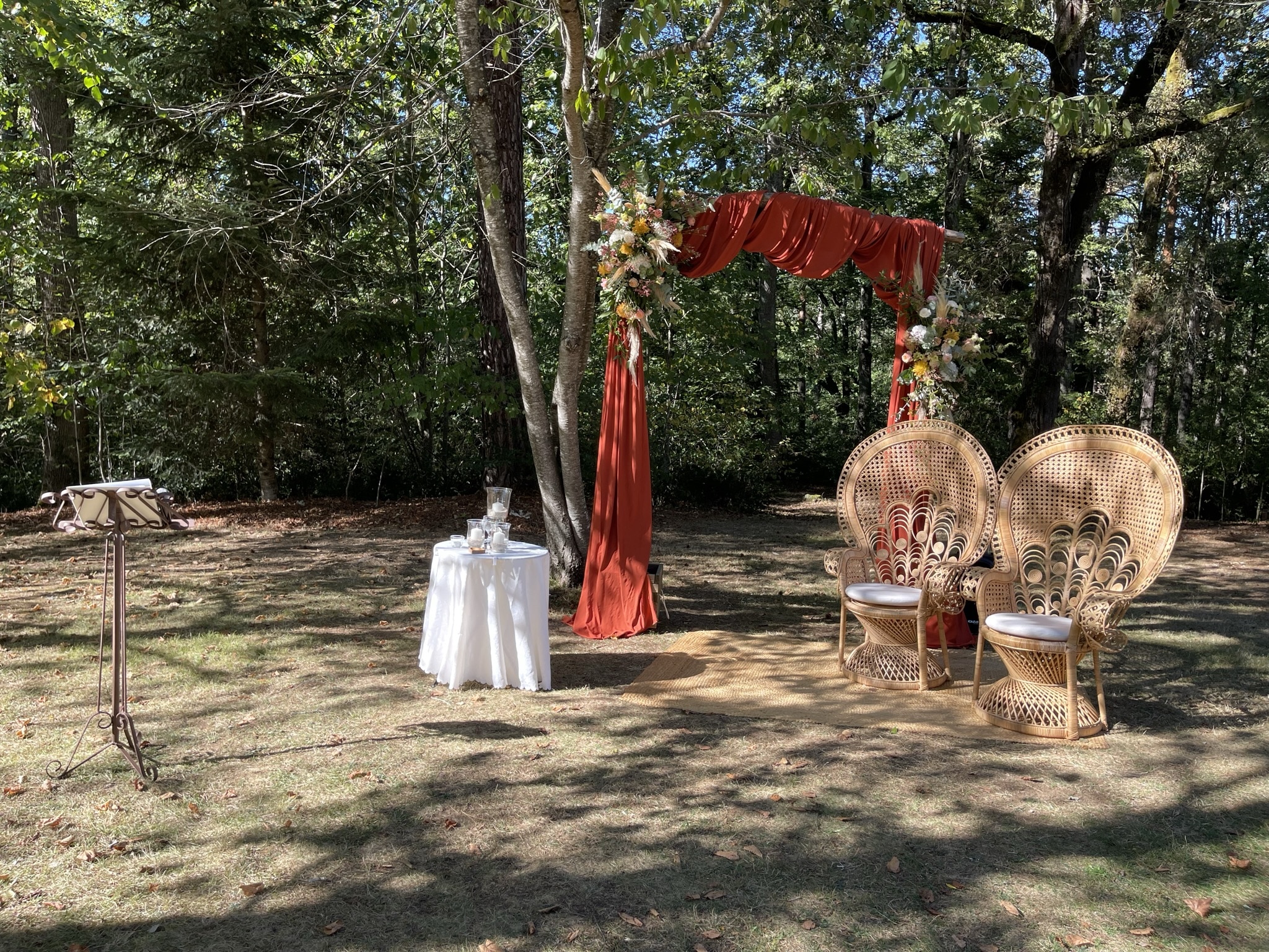 Décoration de mariage en plein air sous un arche en tissu orange avec fleurs, deux chaises en rotin paon, petite table blanche et pupitre en fer forgé.
