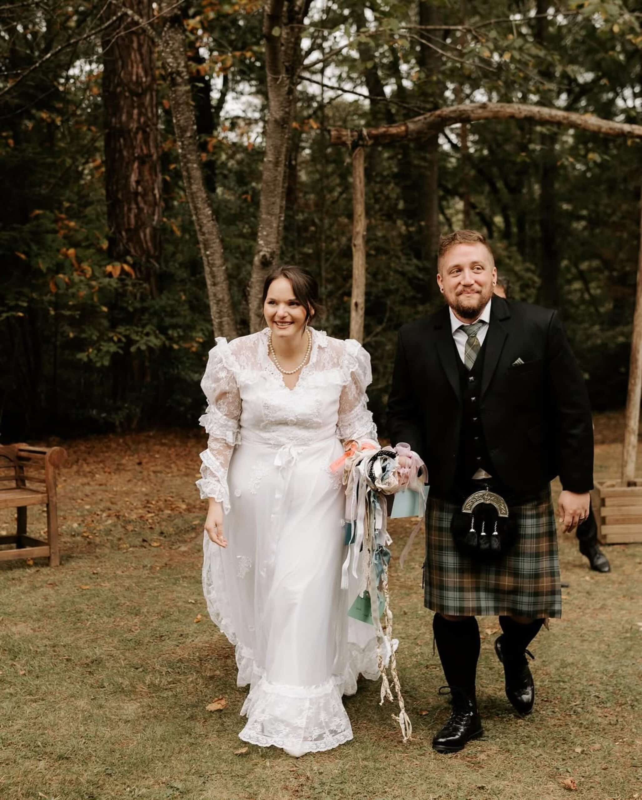 Joyeux couple marié marchant sur l'herbe : la mariée porte une robe en dentelle et le marié un kilt écossais.