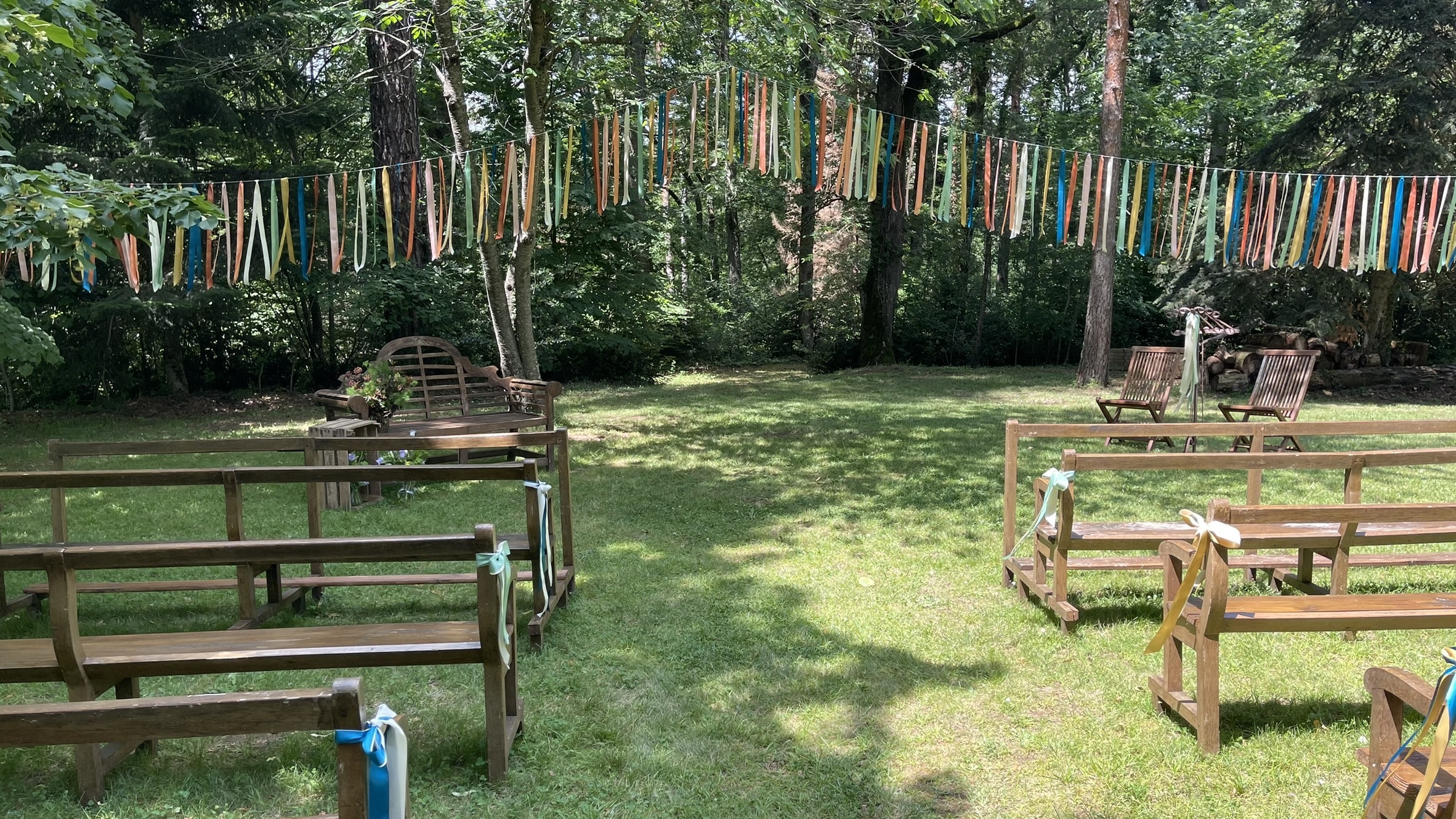 Scène de cérémonie en plein air sous les arbres, décorée de rubans multicolores suspendus et de bancs en bois.