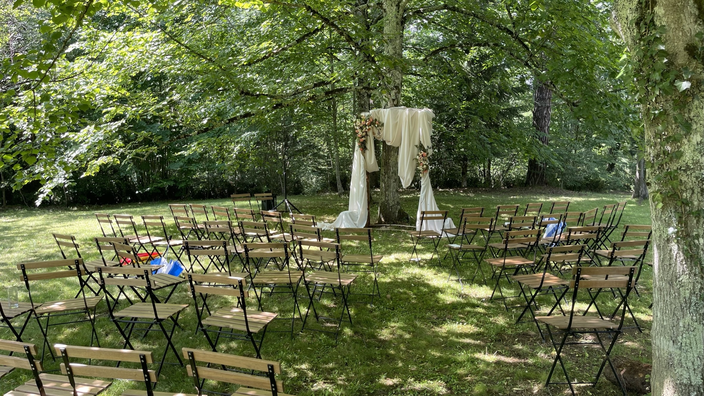 Installation de mariage extérieure sous un grand arbre, avec des chaises pliantes en bois alignées sur la pelouse verte pour une cérémonie.