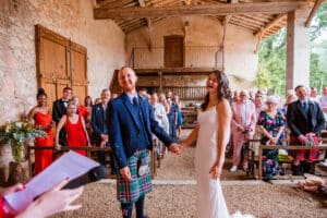 Marié en kilt et mariée riant lors de leur mariage rustique dans une grange en pierre avec des invités souriants en arrière-plan.
