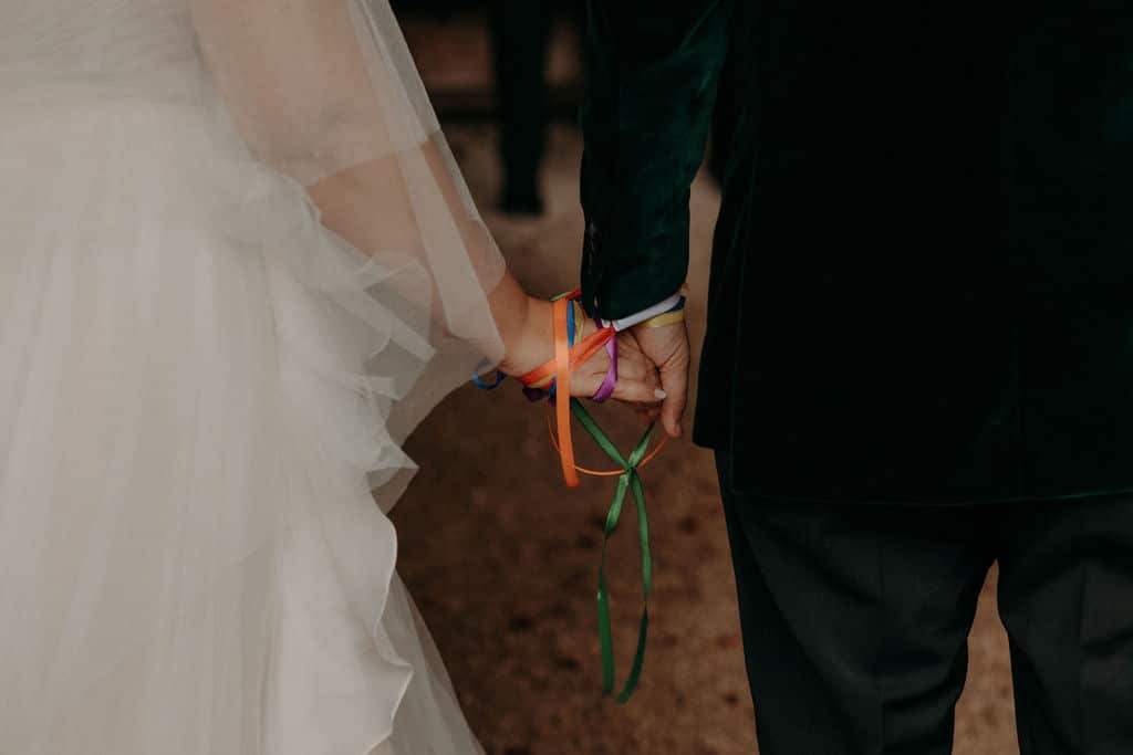 Gros plan sur deux mains jointes, liées par des rubans multicolores (handfasting) lors d'un mariage. Mariée en blanc et marié en velours vert.
