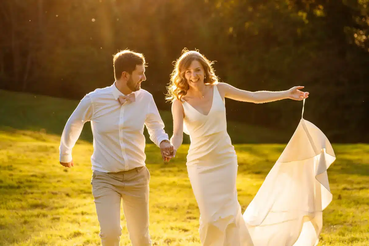 Couple de mariés joyeux au soleil couchant. Mariés joyeux courant main dans la main dans un champ doré. Forte lumière du coucher de soleil illuminant la robe blanche et les sourires.