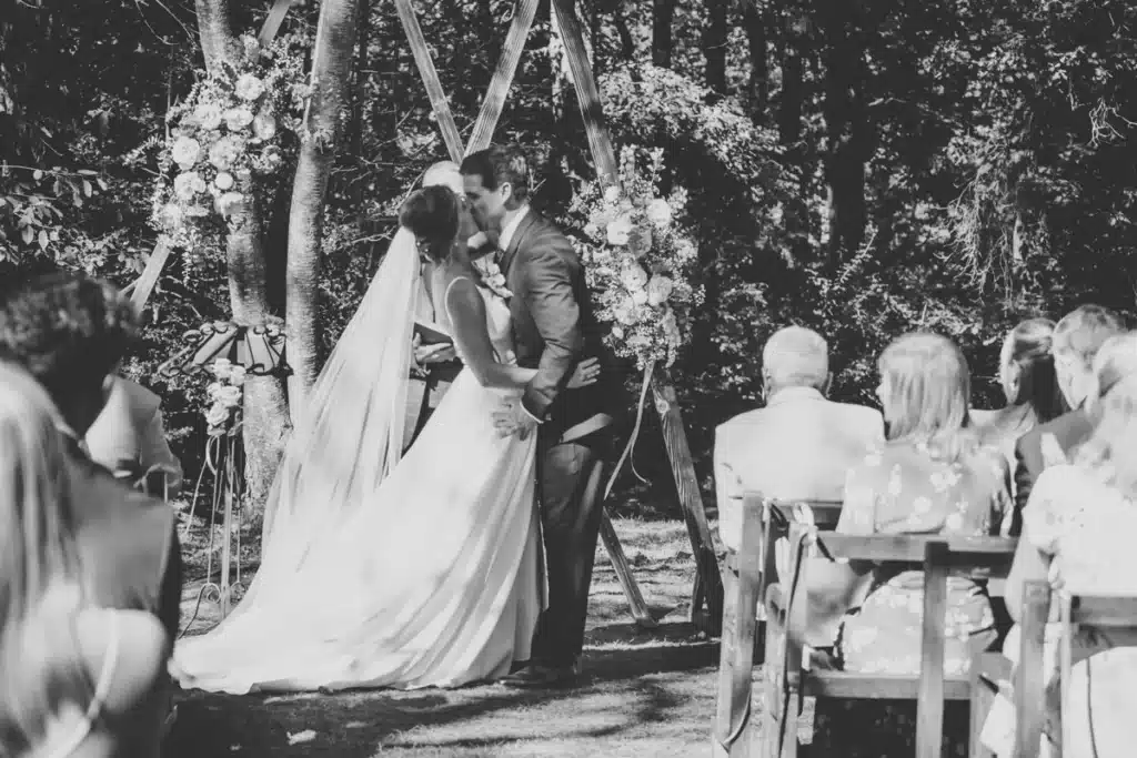 Mariage extérieur : Le baiser des mariés sous l'arche florale Les mariés s'embrassent passionnément devant une arche florale en bois lors de leur cérémonie de mariage en extérieur. Invités au premier plan.