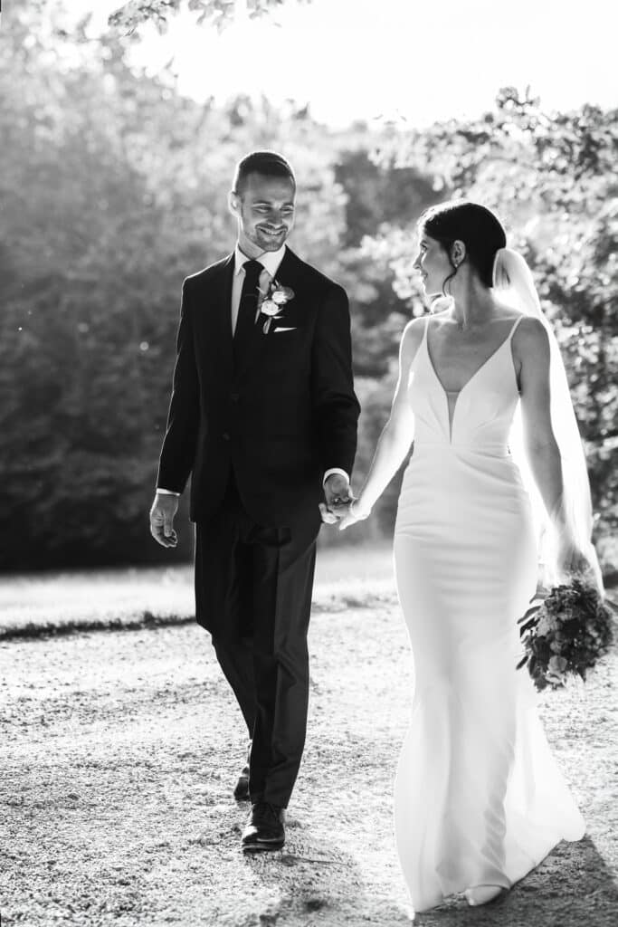 Mariage en Noir et Blanc : Couple Marchant en Plein Air Mariés souriants marchant main dans la main en noir et blanc dans un jardin ensoleillé.