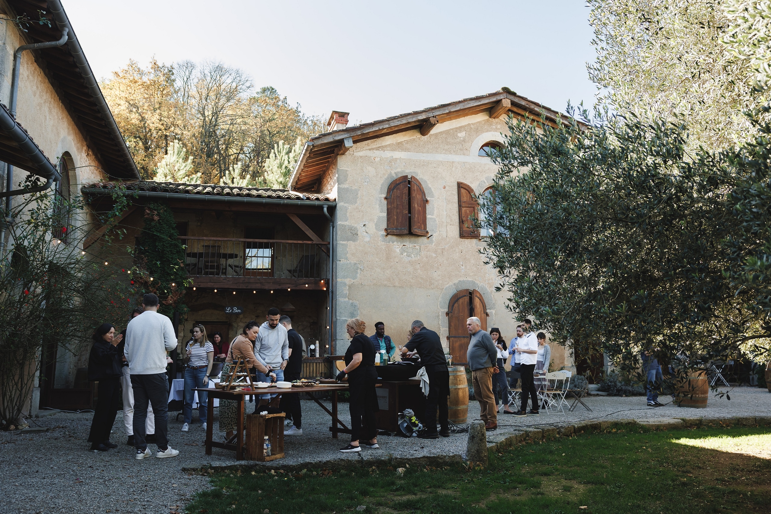 Réception conviviale en plein air dans la cour d'un domaine rustique avec buffet et enseigne Le Bar.