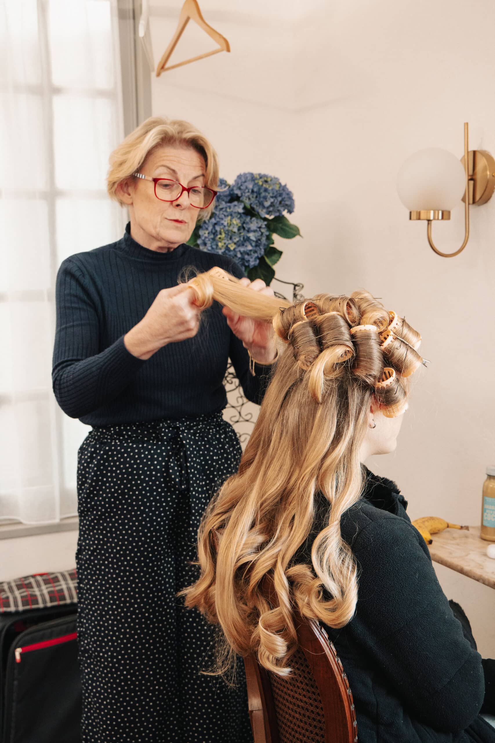 Coiffeuse aux lunettes rouges posant des bigoudis sur les longs cheveux blonds bouclés d'une cliente.