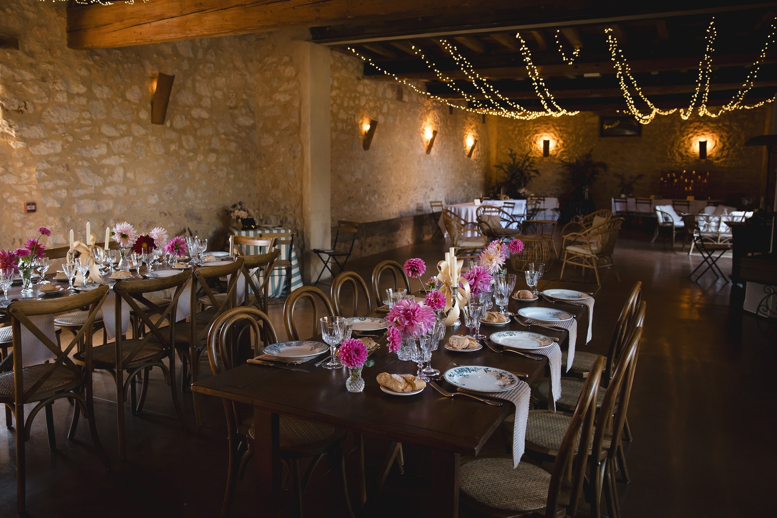 Salle de réception rustique avec murs en pierre, tables dressées, fleurs roses et guirlandes lumineuses chaleureuses.