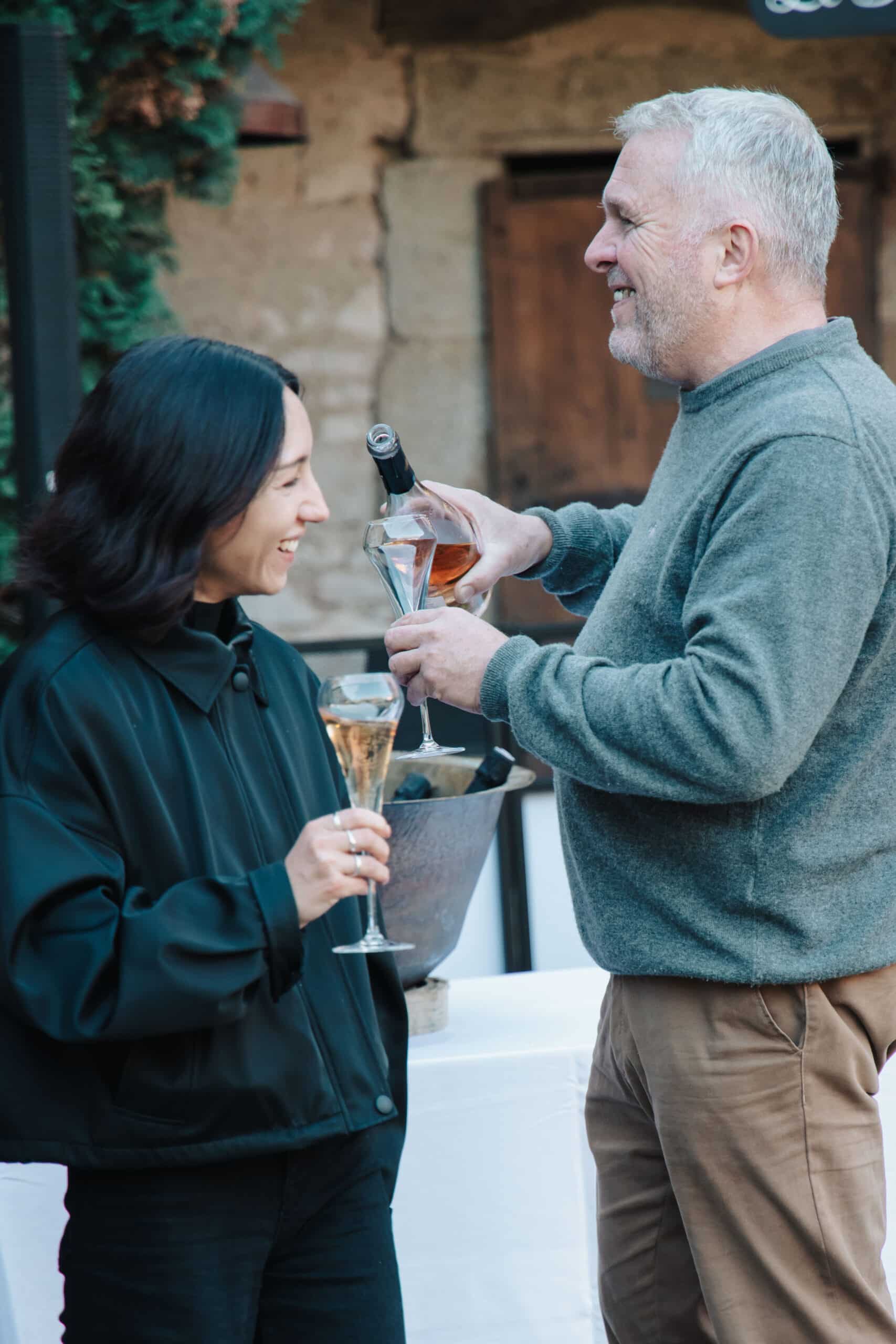 Homme barbu versant du champagne rosé à une femme souriante lors d'un événement festif en plein air.
