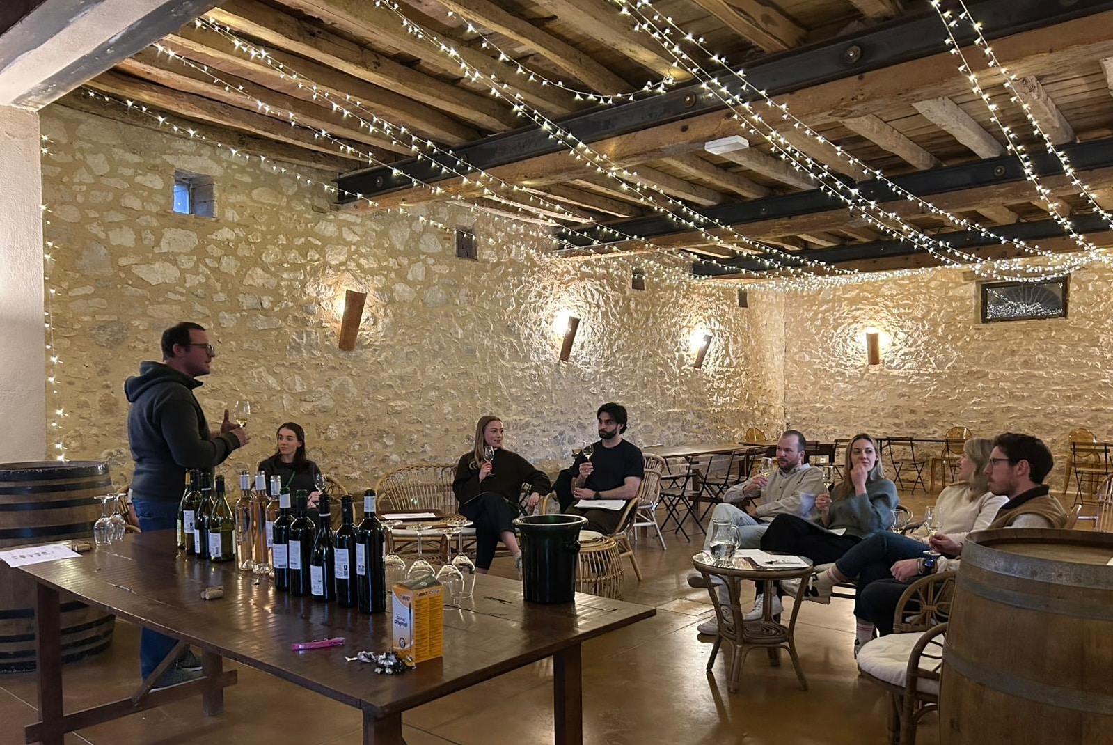 Séance de dégustation de vin conviviale dans un chai rustique avec murs en pierre et guirlandes lumineuses.