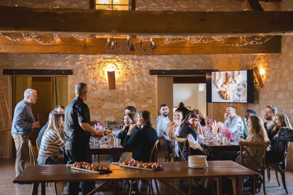 Dîner convivial dans une salle de réception en pierre Groupe convivial dînant dans une salle rustique en pierre avec poutres apparentes, guirlandes et buffet traiteur.