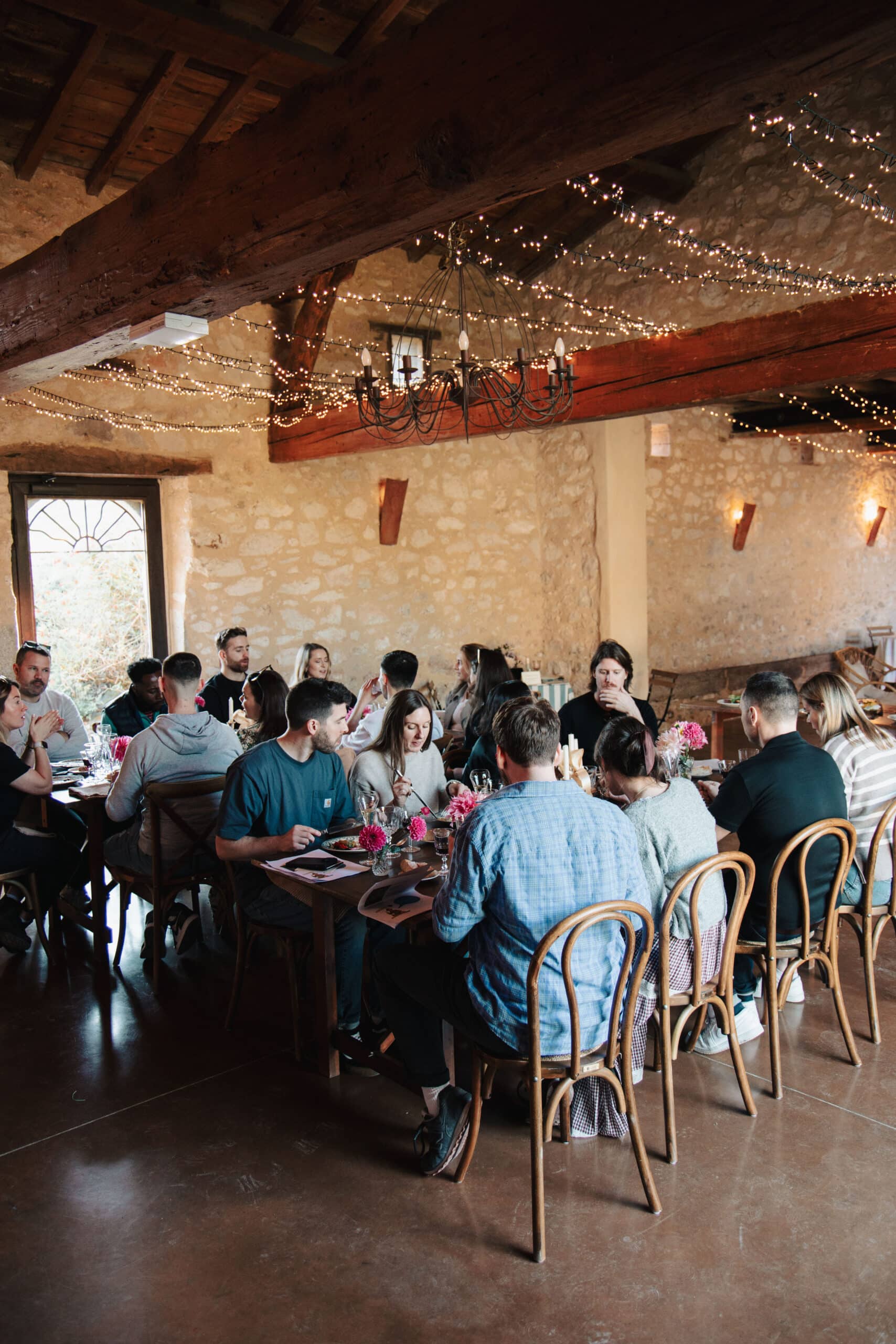 Groupe d'amis dinant à une table rustique sous des guirlandes lumineuses dans une salle aux murs de pierre.
