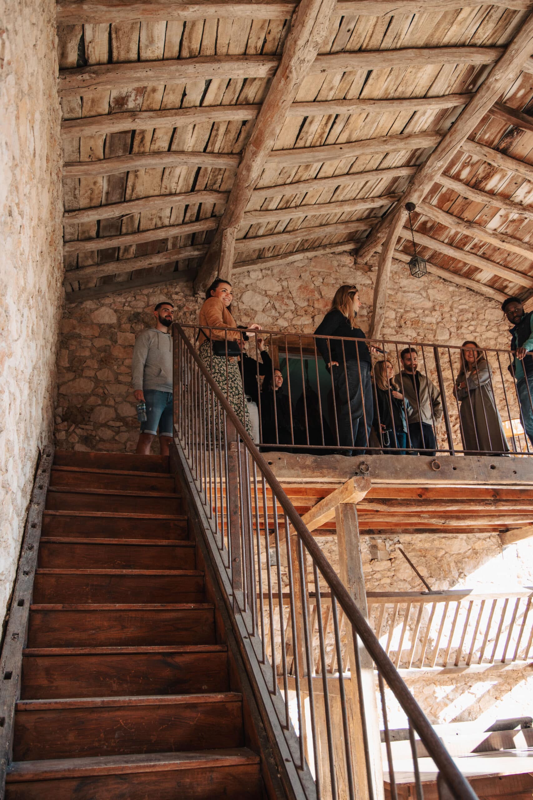 Groupe de visiteurs sur un balcon en bois et pierre dans un bâtiment rustique avec escalier ancien.
